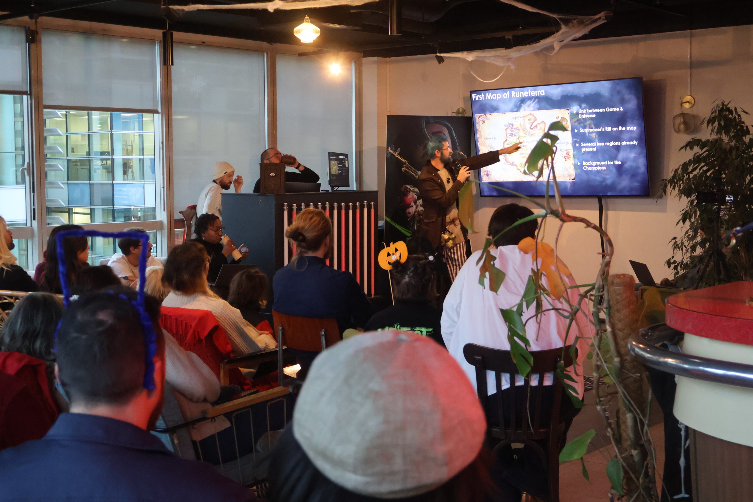 An indoor photo at Fortiche showing a presentation on the lore of Runeterra and the history of League of Legends. An influenceur, cosplaying Jinx, points to a screen. An audience sits in the foreground, and the room is decorated with Halloween-themed items like cobwebs and a pumpkin.