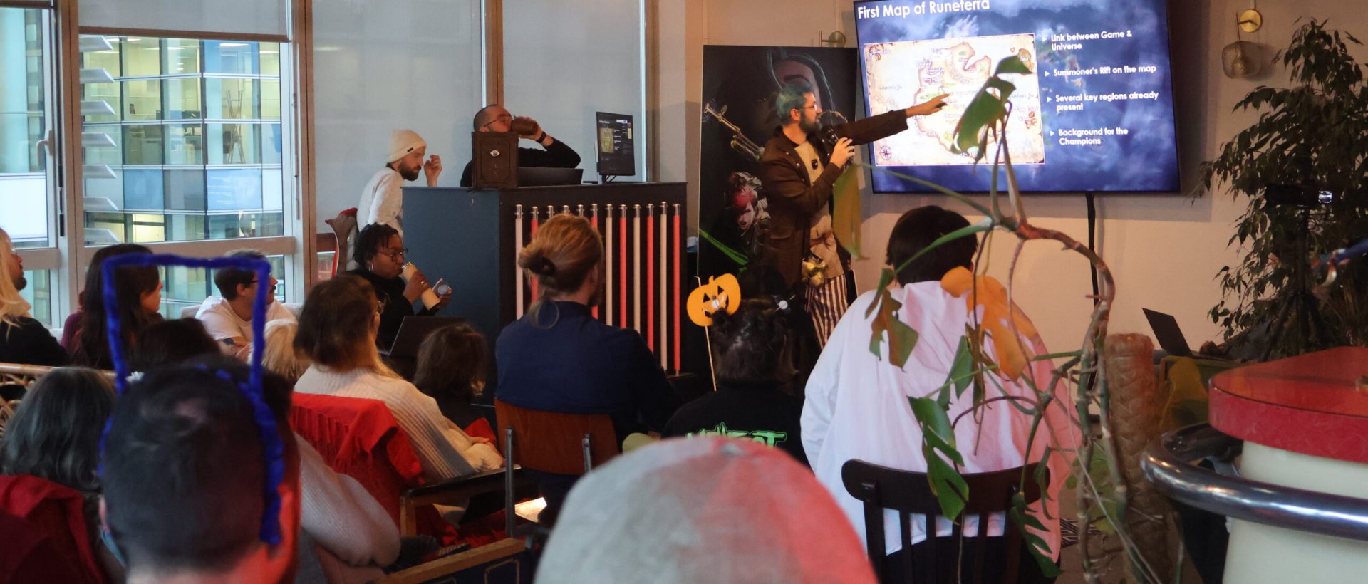 An indoor photo at Fortiche showing a presentation on the lore of Runeterra and the history of League of Legends. An influenceur, cosplaying Jinx, points to a screen. An audience sits in the foreground, and the room is decorated with Halloween-themed items like cobwebs and a pumpkin.